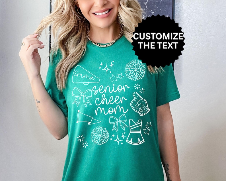 May include: A teal green t-shirt with white graphics, including a megaphone with the name "Emma", bows, stars, pom-poms, a foam finger, and a cheerleader uniform. The text "senior cheer mom" is also printed on the shirt.