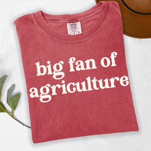 May include: A red t-shirt with the words "big fan of agriculture" printed in white. The shirt is made of a soft material and has a crew neck. The Comfort Colors brand tag is visible at the top.