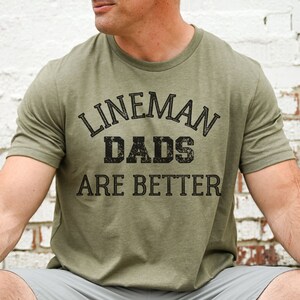 May include: Olive green t-shirt with the text "LINEMAN DADS ARE BETTER" in black, distressed lettering. The shirt has a crew neck and short sleeves. The person is wearing gray shorts.