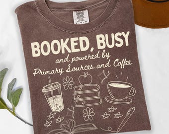 May include: A brown t-shirt with the text "BOOKED, BUSY and powered by Primary Sources and Coffee." The shirt features white illustrations of a drink, books, flowers, an apple, a coffee cup, and a pen. A brown hat is partially visible.