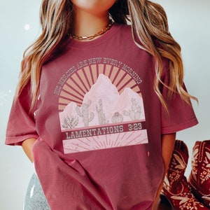 May include: A maroon t-shirt with a graphic design of a desert landscape with cacti and a sunrise. The text "His mercies are new every morning" and "Lamentations 3:23" are printed on the shirt.
