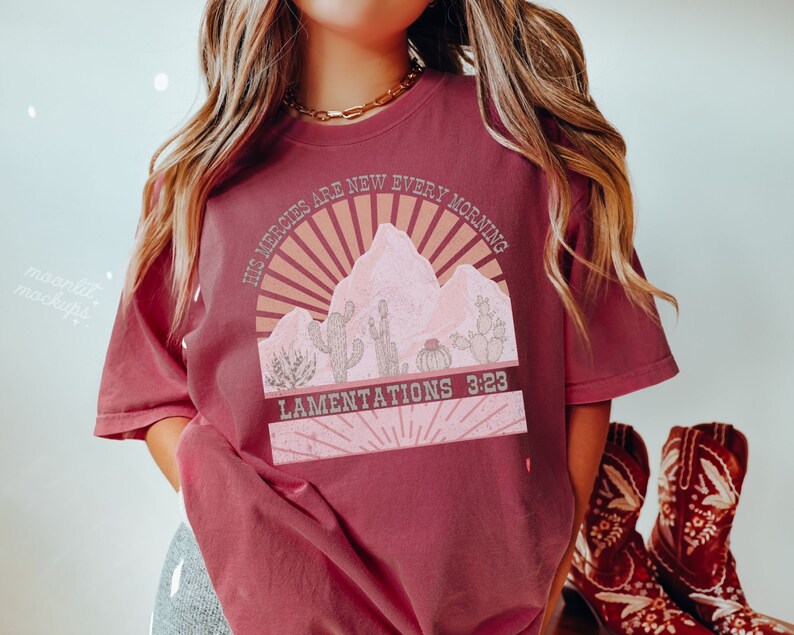 May include: A maroon t-shirt with a graphic design of a desert landscape with cacti and a sunrise. The text "His mercies are new every morning" and "Lamentations 3:23" are printed on the shirt.