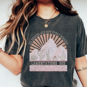 May include: A dark gray t-shirt with a vintage-style graphic of a mountain range with a sunrise and cacti. The text "His mercies are new every morning" and "Lamentations 3:23" are printed on the shirt.