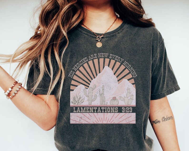 May include: A dark gray t-shirt with a vintage-style graphic of a mountain range with a sunrise and cacti. The text "His mercies are new every morning" and "Lamentations 3:23" are printed on the shirt.