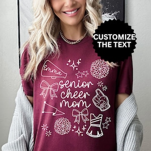 May include: Burgundy t-shirt with white graphic elements and the text "senior cheer mom." The design includes a megaphone, bows, stars, pom-poms, and a cheerleader uniform. The text "Customize the Text" is also visible.