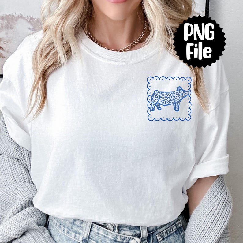 May include: White t-shirt with a blue pig illustration inside a scalloped square. The pig has a heart pattern. The shirt is a casual style.