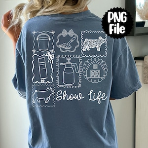 May include: A blue t-shirt with white line drawings of farm-related items, including a bucket, brush, pig, spray bottle, and barn. The words "Show Life" are written in script below the drawings. A black and white "PNG File" graphic is in the upper right corner.