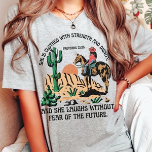 May include: A heather gray t-shirt featuring a desert scene with a cactus, sun, and a person riding a horse. The shirt includes the text "SHE IS CLOTHED WITH STRENGTH AND DIGNITY" and "AND SHE LAUGHS WITHOUT FEAR OF THE FUTURE."