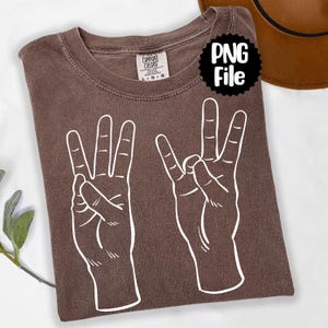 Könnte beinhalten: Ein braunes T-Shirt mit einer weißen Grafik von zwei Händen, die das "Drei"-Zeichen machen. Das Shirt hat einen Rundhalsausschnitt und ein Etikett mit der Aufschrift "Comfort Colors". Ein schwarz-weißer "PNG File"-Aufkleber befindet sich auf dem Shirt.