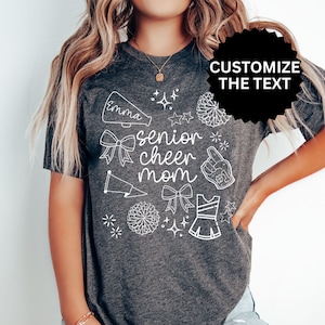 May include: Dark gray t-shirt with white graphic text and illustrations. The shirt says "senior cheer mom" with a name above the text. Cheerleading-themed illustrations include bows, stars, and a megaphone.
