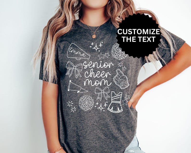 May include: Dark gray t-shirt with white graphic text and illustrations. The shirt says "senior cheer mom" with a name above the text. Cheerleading-themed illustrations include bows, stars, and a megaphone.