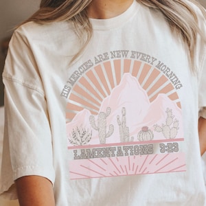 May include: A white t-shirt with a pink and brown graphic design featuring a mountain range with a sunrise and cacti. The text "His mercies are new every morning" and "Lamentations 3:23" are printed on the shirt.