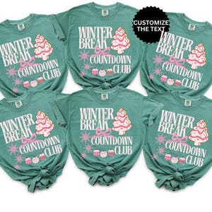 May include: Six teal t-shirts with the text "Winter Break Countdown Club" in white, with a pink Christmas tree and ornaments design. The shirts have short sleeves and are knotted at the bottom.