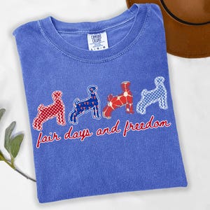 May include: A blue Comfort Colors t-shirt featuring four dog silhouettes in red, white, and blue patterns. The text "fair days and freedom" is written in red script across the bottom of the shirt.