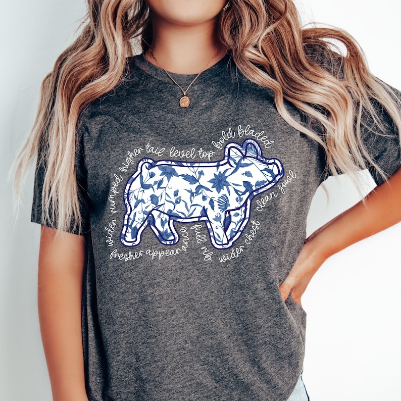 May include: Dark gray t-shirt featuring a pig illustration filled with blue floral patterns. The pig design is outlined in white, with text around the pig that reads "wider rumped higher tail level top bold bladed wider chest clean jowl".