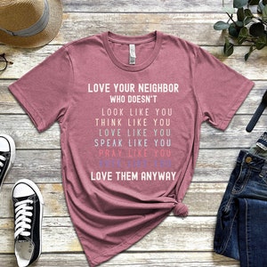 Op de afbeelding: Een roze heather t-shirt met de tekst "LOVE YOUR NEIGHBOR WHO DOESN'T LOOK LIKE YOU THINK LIKE YOU LOVE LIKE YOU SPEAK LIKE YOU PRAY LIKE YOU LOVE THEM ANYWAY" in wit gedrukt.