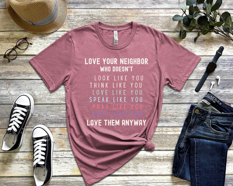 Op de afbeelding: Een roze heather t-shirt met de tekst "LOVE YOUR NEIGHBOR WHO DOESN'T LOOK LIKE YOU THINK LIKE YOU LOVE LIKE YOU SPEAK LIKE YOU PRAY LIKE YOU LOVE THEM ANYWAY" in wit gedrukt.