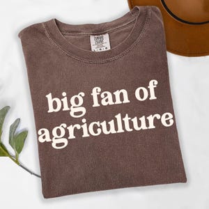 May include: A brown t-shirt with the text "big fan of agriculture" in white, with a Comfort Colors tag. A brown hat is partially visible in the upper right corner. The shirt is folded and lies on a white surface.