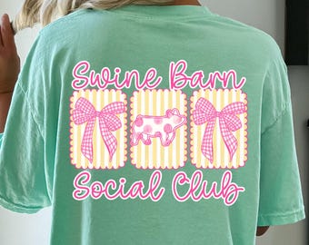 May include: A light green t-shirt with the words "Swine Barn Social Club" in pink script. The design includes three rectangular images with a pink bow, a pig, and another bow. The pig is white with pink spots.