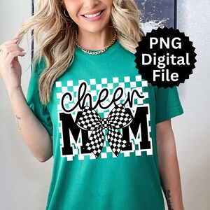 Puede incluir: Camiseta verde azulado con un gr&aacute;fico que dice "cheer MOM" en blanco y negro. El dise&ntilde;o incluye un lazo a cuadros y un fondo cuadrado a cuadros. Un logotipo de archivo digital PNG negro est&aacute; en la esquina superior derecha.