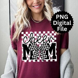Puede incluir: Camiseta burdeos con un dise&ntilde;o a cuadros blancos y negros. La camiseta presenta las palabras "cheer MOM" en una fuente audaz y estilizada, con un gr&aacute;fico de lazo a cuadros. Tambi&eacute;n hay un archivo digital PNG disponible.
