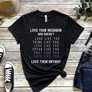 Op de afbeelding: Zwart T-shirt met de tekst "LOVE YOUR NEIGHBOR WHO DOESN'T LOOK LIKE YOU THINK LIKE YOU LOVE LIKE YOU SPEAK LIKE YOU PRAY LIKE YOU VOTE LIKE YOU LOVE THEM ANYWAY"