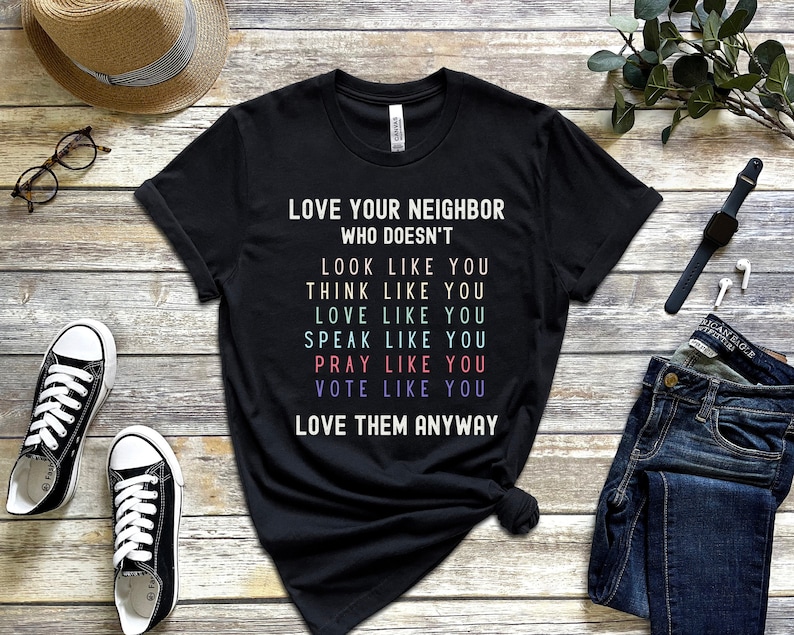 Op de afbeelding: Zwart T-shirt met de tekst "LOVE YOUR NEIGHBOR WHO DOESN'T LOOK LIKE YOU THINK LIKE YOU LOVE LIKE YOU SPEAK LIKE YOU PRAY LIKE YOU VOTE LIKE YOU LOVE THEM ANYWAY"