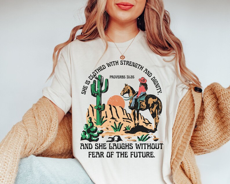 May include: Cream-colored t-shirt featuring a desert scene with a cactus, a sunset, and a person riding a horse. The shirt has the text "SHE IS CLOTHED WITH STRENGTH AND DIGNITY" and "AND SHE LAUGHS WITHOUT FEAR OF THE FUTURE."