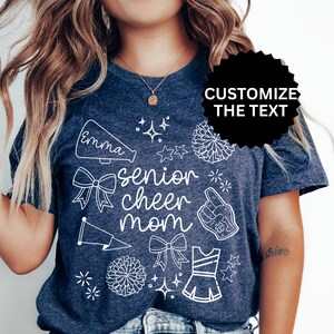 May include: Navy blue t-shirt with white graphic elements, including a megaphone with the name "Emma", bows, pom-poms, and the words "senior cheer mom". The shirt also features a cheerleading uniform and a hand with the number one.