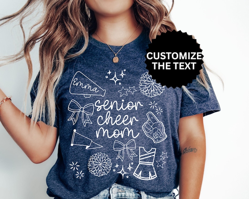 May include: Navy blue t-shirt with white graphic elements, including a megaphone with the name "Emma", bows, pom-poms, and the words "senior cheer mom". The shirt also features a cheerleading uniform and a hand with the number one.