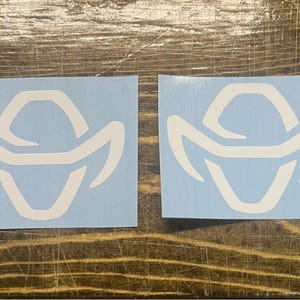 May include: Two white vinyl stickers with a stylized cowboy hat design on a light blue background. The stickers are rectangular and feature a bold, geometric outline of a cowboy hat. The stickers are placed on a wooden surface.