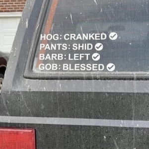 以下が含まれることがあります： 車両の後部窓に貼られたステッカーで、「HOG: CRANKED」、「PANTS: SHID」、「BARB: LEFT」、「GOB: BLESSED」の各フレーズがチェックマーク付きで表示されています。車両は濃い色で、ほこりっぽく見えます。