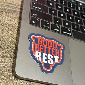 Puede incluir: Una pegatina con las palabras "GOOD BETTER BEST" en un dise&ntilde;o rojo y blanco, delineado en azul. La pegatina est&aacute; colocada en un port&aacute;til plateado, con el teclado visible.