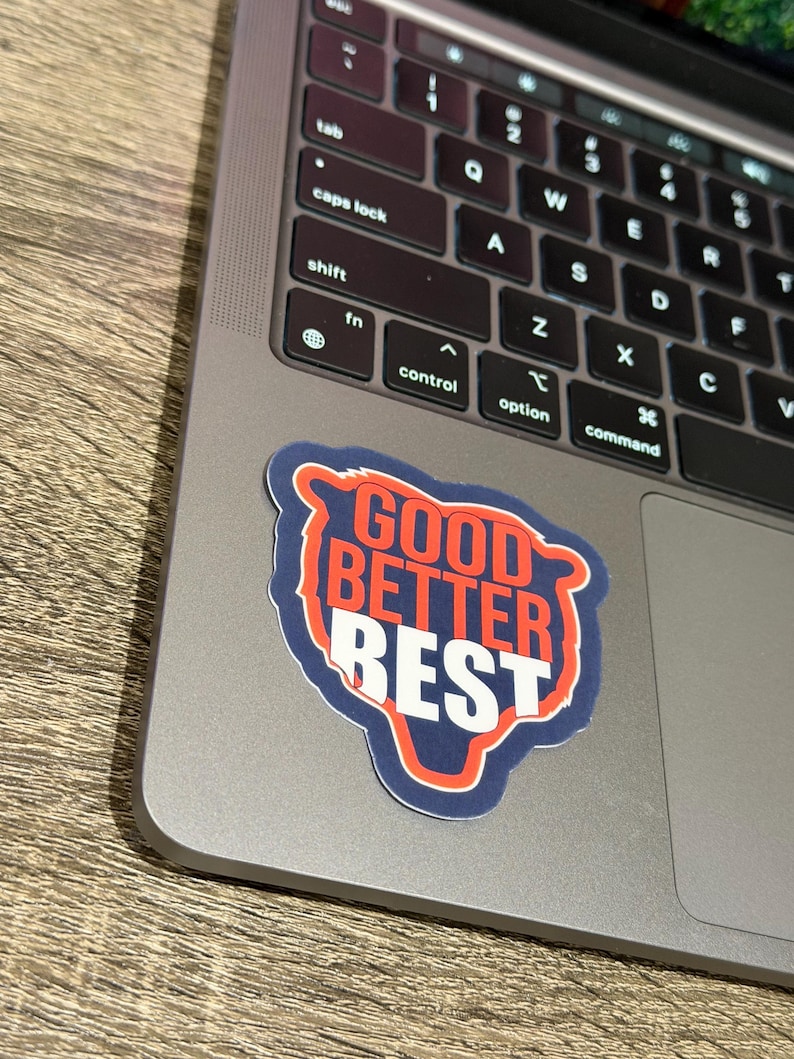 Puede incluir: Una pegatina con las palabras "GOOD BETTER BEST" en un dise&ntilde;o rojo y blanco, delineado en azul. La pegatina est&aacute; colocada en un port&aacute;til plateado, con el teclado visible.