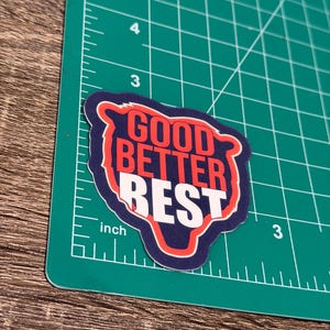 Puede incluir: Una pegatina con las palabras "GOOD BETTER REST" en rojo y blanco, delineadas en blanco, sobre un fondo azul marino. La pegatina est&aacute; sobre una esterilla de corte verde con medidas en pulgadas.