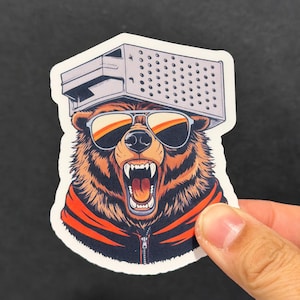 May include: A sticker featuring a cartoon bear with sunglasses and a grater on its head, roaring with its mouth open. The bear is wearing a red and black jacket. The sticker has a white border.