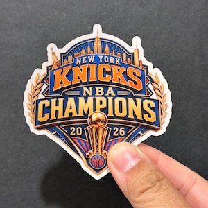 May include: A die-cut sticker featuring a New York Knicks NBA Champions 2026 graphic. The design includes a gold trophy, a blue and orange color scheme, a skyline silhouette, and laurel leaf accents, all set against a shield-shaped background.