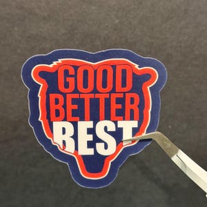 Puede incluir: Una pegatina con las palabras "GOOD BETTER BEST" en rojo y blanco sobre un fondo azul en forma de oso, con un borde rojo. Unas pinzas sujetan la pegatina, lista para ser aplicada.