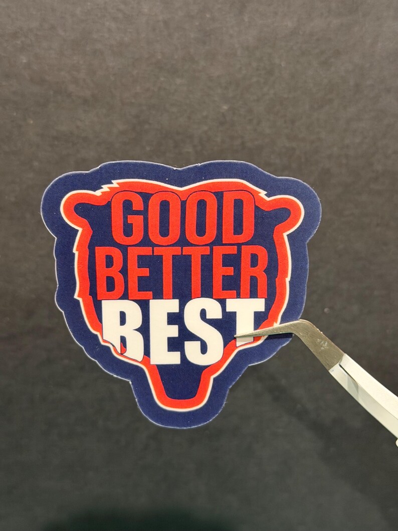 Puede incluir: Una pegatina con las palabras "GOOD BETTER BEST" en rojo y blanco sobre un fondo azul en forma de oso, con un borde rojo. Unas pinzas sujetan la pegatina, lista para ser aplicada.