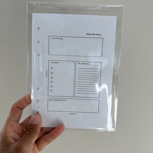 Könnte beinhalten: Ein weißes Daily Devotion-Planereinlage mit schwarzem Text, gehalten in einer transparenten Plastikhülle. Der Einsatz enthält Abschnitte für "Vers des Tages", "Schlüsselpunkte" und "Lebensanwendung."