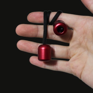 May include: A pair of red metal fidget toys with black string attached. The toys are hanging from a hand.
