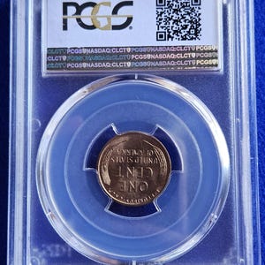 Puede incluir: Una moneda de un centavo de cobre de los Estados Unidos, encapsulada en un soporte de plástico transparente. La moneda está centrada dentro de un soporte teñido de azul, con el texto "ONE CENT" y "UNITED STATES OF AMERICA" visible. El soporte tiene el logotipo "PCGS".