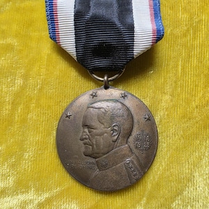 May include: A bronze-toned circular military medal featuring a profile relief of General John J. Pershing, surrounded by five stars and the dates 1918 and 1919. The medal hangs from a striped ribbon in black, white, red, and blue colors.