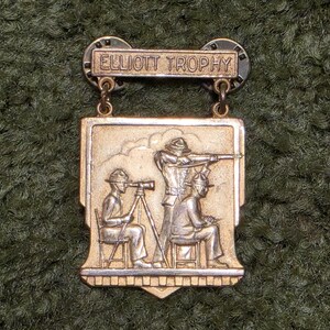 May include: A metallic gold-toned pin featuring the text ELLIOTT TROPHY on a rectangular top bar. The main shield-shaped section depicts a relief scene of three figures in hats, with one person using a spotting scope on a tripod and another aiming a rifle.
