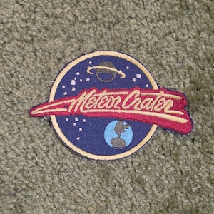 Könnte beinhalten: Ein kreisförmiger gestickter Aufnäher mit dunkelblauem Hintergrund, weißen Sternen, einem braunen Planeten mit Ringen und einer hellblauen Weltkugel. Ein rotes Banner mit Goldrand überlagert die Mitte und zeigt den Text Meteor Crater in Goldschrift.