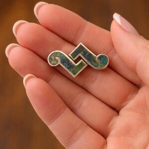 May include: Silver brooch with an abstract design. The brooch has a geometric shape with a central square and curved edges, inlaid with a mosaic of blue and green stones. The brooch is held in a hand.