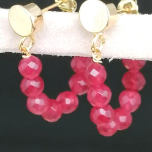 Puede incluir: Pendientes de botón dorados con un poste circular y un diseño colgante en forma de U. La parte colgante está hecha de cuentas facetadas de color rosa brillante. Los pendientes se muestran sobre un fondo blanco.