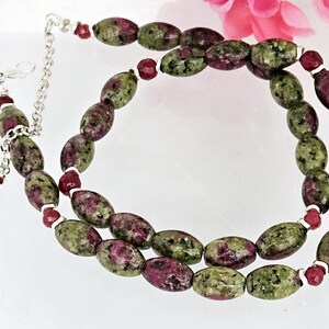 May include: A necklace with oval beads in green and burgundy, with smaller red beads. The necklace has a silver chain and clasp, with a matching dangling bead. The jewellery is displayed on a white surface.