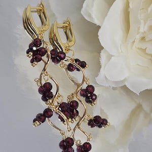 May include: Gold-coloured earrings featuring clusters of dark red beads, reminiscent of grapes, suspended from a curved vine design. The earrings have a lever back closure. The background is a soft, blurred white.