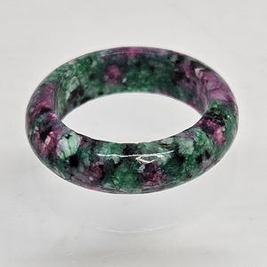 May include: A gemstone ring with a polished finish, displaying a marbled effect. The ring combines green, black, and deep pink colours. The ring is a solid band, highlighting the natural patterns of the stone. The ring is a piece of jewellery.